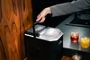 A hand holds the handle of a portable ice maker while two drinks sit on a counter. Podgląd zdjęcia produktu