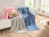 Several blankets are folded and draped on a white sofa in a well-lit room with a large window. Produktbild-Vorschau
