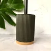 A toilet brush with a ribbed black body and a wooden base, placed on a marble-patterned surface with a large green leaf in the background. Anteprima dell’immagine prodotto