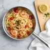 A close-up overhead view of a stainless steel pan filled with spaghetti, shrimp, and cherry tomatoes, with a lemon and parsley on a wooden board to the side. Náhled obrázku produktu