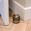 A close-up view of a silver door stopper with black rubber rings on a light wood floor, next to a white baseboard and a white door. Náhled obrázku produktu
