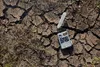 A camera with a strap lies on cracked, dry earth with some sparse vegetation. Náhled obrázku produktu