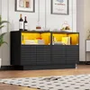 A modern black sideboard with yellow interior lighting, displaying books, a radio, wine bottles, and decorative items. Produktbild-Vorschau