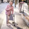 An elderly woman is walking on a sidewalk with a cane that has a tripod base. Produktbild-Vorschau