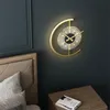 A modern gold wall clock with warm white light is mounted on a dark teal wall above a wooden bedside table with a book on it. A portion of a bed with a textured headboard and pillows is visible on the left. Produktbild-Vorschau