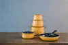 A set of orange pots and a frying pan are arranged on a wooden table in front of a gray wall. Podgląd zdjęcia produktu