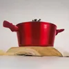A red pot with a lid sits on a wooden surface with some pasta. Náhled obrázku produktu