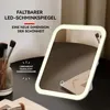 A makeup mirror with LED lighting is shown on a dark surface, with makeup brushes in a jar in the background. Produktbild-Vorschau