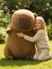 A young girl is hugging a large plush capybara toy in a grassy outdoor setting. Produktbild-Vorschau
