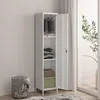 A tall, narrow white metal locker with its door open, revealing shelves with folded clothes, a basket, and a green box. A wire laundry basket with clothes is to the left. The scene is set against a light gray wall and a window with curtains and a plant. Produktbild-Vorschau