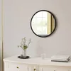 A round black mirror hangs on a light gray wall above a white dresser. A vase with white flowers sits on the dresser to the left of a glass of water and a stack of books. Produktbild-Vorschau
