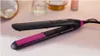A black and pink hair straightener is placed on a light pink surface, with blurred cosmetic items in the background. Voir les images du produit