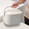 A hand holding a white trash bin with a gray handle. Voir les images du produit