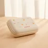 A small, square-shaped beige case with a pattern of small orange fruits and green leaves, placed on a wooden surface. The background shows soft, diffused light filtering through curtains. Produktbild-Vorschau