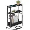A narrow black side table with integrated charging ports and shelves, shown against a white background. It features a tablet, a digital clock, and books on its shelves. Náhled obrázku produktu