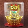 A golden Maneki Neko cat figurine with a solar-powered waving paw, displayed in a clear plastic case on a red base, set against a textured brown background. Produktbild-Vorschau
