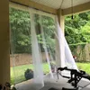 A covered patio or porch area with sheer white curtains blowing in the breeze, overlooking a backyard with trees and a fence. A piece of exercise equipment is visible in the foreground. Voir les images du produit
