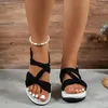 Frau trägt schwarze Keilsandalen mit geknoteten Riemen und einen weißen Perlenfußkettchen, stehend auf grauem Boden Anteprima dell’immagine prodotto