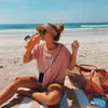 A young woman is sitting on a beach towel on the sand, drinking from an orange water bottle. She is wearing a pink t-shirt and a straw visor. A straw beach bag and a book are next to her. Produktbild-Vorschau