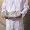 A person wearing a white lab coat holds up a white shoe, showing the sole. Produktbild-Vorschau
