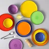 A collection of colorful ceramic plates and bowls arranged on a light gray surface. Produktbild-Vorschau
