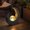 A solar-powered crescent moon lamp with a crackled glass ball sits on a tiled patio at dusk, with a hammock and potted plants in the background. Anteprima dell’immagine prodotto