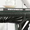 A close-up view of a black rattan side table with a glass top, a white cup and saucer on top, and a book with the title 'History of the Greatest Places' visible. Produktbild-Vorschau