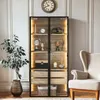 A tall, narrow display cabinet with glass doors and wooden shelves, filled with decorative items, stands next to an armchair in a well-lit room. Anteprima dell’immagine prodotto