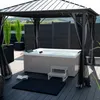 An outdoor whirlpool is set up on a wooden deck under a gazebo, with lush green plants and a dark fence in the background. Podgląd zdjęcia produktu