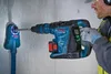 A person is using a cordless rotary hammer drill to drill a hole in a concrete wall, with a dust extraction system attached. Produktbild-Vorschau
