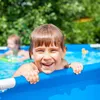 A young child is smiling and holding onto the edge of a blue swimming pool. Produktbild-Vorschau