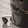 A close-up shot of a black metal fire pit with cut-out lettering and a wheel attached to its base, resting on a paved surface. Produktbild-Vorschau