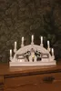 A white LED wooden silhouette of houses with candles on a wooden dresser, with a patterned wallpaper background. Produktbild-Vorschau