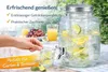 A hand is pouring a drink from a glass dispenser into a glass, with fruit and other drinks in the background, set outdoors on a table. Produktbild-Vorschau