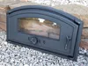 A black cast iron stove door with a glass window rests on a bed of grey gravel, with large stones in the background. Produktbild-Vorschau
