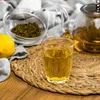 A glass of tea, a lemon, and dried tea leaves are arranged on a woven mat. Produktbild-Vorschau