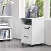 A white rolling filing cabinet with two drawers and an open shelf, placed next to a window with greenery visible outside. A magazine rack with magazines and a vase with flowers are on top of the cabinet. Produktbild-Vorschau
