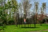 A child is jumping on a black trampoline in a grassy backyard with trees in the background. Náhled obrázku produktu