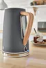 A close-up shot of a grey electric kettle with a wooden handle, placed on a wooden countertop. In the background, a jar of jam and some toast are visible, suggesting a breakfast setting. Produktbild-Vorschau