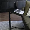 A laptop is placed on a black, adjustable folding table next to a sofa. Náhľad obrázku produktu