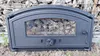 A black cast iron oven door with a glass window filled with stones, placed on the ground in front of a stone wall. Produktbild-Vorschau