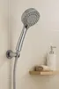 A chrome hand shower head with water spraying from it, mounted on a tiled wall. A small shelf with a folded towel and a soap dispenser is visible to the right. Produktbild-Vorschau