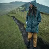 A woman wearing a hat and jacket walks on a muddy path in a grassy, hilly landscape. Produktbild-Vorschau