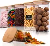 A collection of clear storage jars with bamboo lids, filled with various food items like marshmallows, candies, coffee beans, crackers, walnuts, and pasta. One jar has a chalk-like label indicating its capacity. Náhľad obrázku produktu