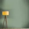 A floor lamp with a yellow shade stands on a wooden floor against a green wall. Produktbild-Vorschau