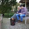 A couple sits on a bench next to a fire pit, holding mugs. Produktbild-Vorschau