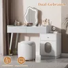 A white vanity table with a mirror, stool, and drawers, set in a room with curtains and a plant. Podgląd zdjęcia produktu