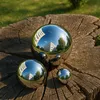 Three polished stainless steel garden balls of different sizes are placed on a weathered wooden stump, with green grass in the background. Náhled obrázku produktu