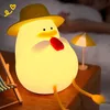 A cute duck-shaped night light sits on a wooden table next to a miniature beach chair and umbrella, with a person sleeping in the background. Produktbild-Vorschau