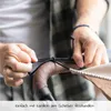 A person's hands are shown tying a cord around a stroller handle, with a beige sunshade visible. Produktbild-Vorschau